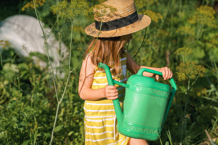 Farmer girl in yellow dress. Little vegetables gardener farming in garden. Big green watering can water fresh cucumbers seedlings. Harvest help work. Cultivation healthy organic food, countryside.の写真素材