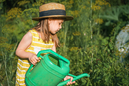 Farmer girl in yellow dress. Little vegetables gardener farming in garden. Big green watering can water fresh cucumbers seedlings. Harvest help work. Cultivation healthy organic food, countryside.の写真素材