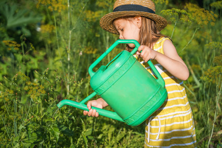 Farmer girl in yellow dress. Little vegetables gardener farming in garden. Big green watering can water fresh cucumbers seedlings. Harvest help work. Cultivation healthy organic food, countryside.の写真素材