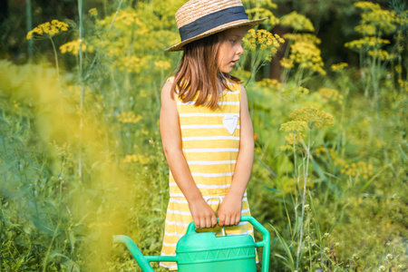 Farmer girl in yellow dress. Little vegetables gardener farming in garden. Big green watering can water fresh cucumbers seedlings. Harvest help work. Cultivation healthy organic food, countryside.の写真素材