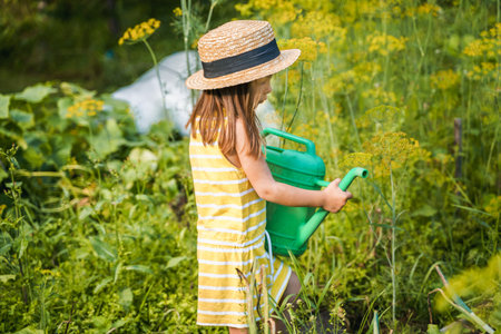 Farmer girl in yellow dress. Little vegetables gardener farming in garden. Big green watering can water fresh cucumbers seedlings. Harvest help work. Cultivation healthy organic food, countryside.の写真素材