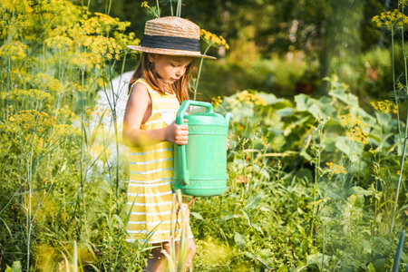 Farmer girl in yellow dress. Little vegetables gardener farming in garden. Big green watering can water fresh cucumbers seedlings. Harvest help work. Cultivation healthy organic food, countryside.の写真素材