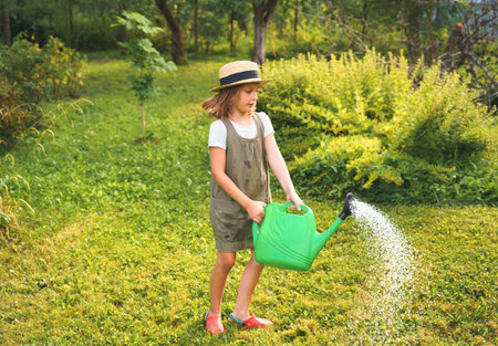Farmer girl in summer straw hat. Little gardener farming spinning, having fun in garden with big green watering can water fresh grass. Harvest help work. Cultivation healthy organic food, countryside.の写真素材