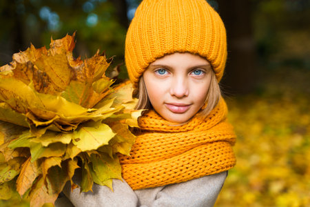 Beautiful little girl in bright yellow orange autumn warm knitted hat and scarf snood with bouquet of maple leaves,foliage walking in forest. Fun playing in the park. Outdoor family activity in nature.の写真素材