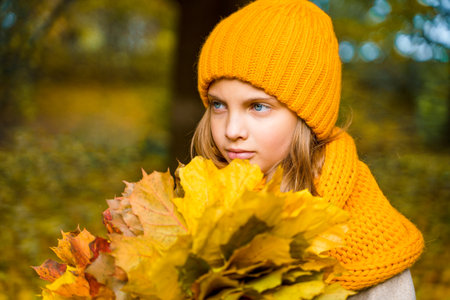 Beautiful little girl in bright yellow orange autumn warm knitted hat and scarf snood with bouquet of maple leaves,foliage walking in forest. Fun playing in the park. Outdoor family activity in nature.の写真素材