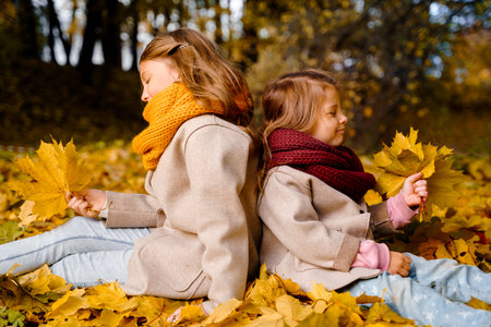 Beautiful little girls in bright yellow red orange autumn warm knitted scarf snood with head wreath of maple leaves,foliage walking in forest. Fun playing in the park. Outdoor family activity in nature.の写真素材
