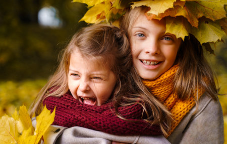 Beautiful little girls in bright yellow red orange autumn warm knitted scarf snood with head wreath of maple leaves,foliage walking in forest. Fun playing in the park. Outdoor family activity in nature.の写真素材