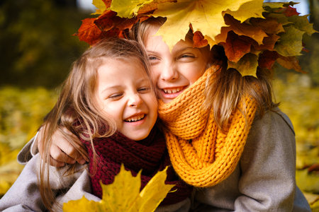 Beautiful little girls in bright yellow red orange autumn warm knitted scarf snood with head wreath of maple leaves,foliage walking in forest. Fun playing in the park. Outdoor family activity in nature.の写真素材