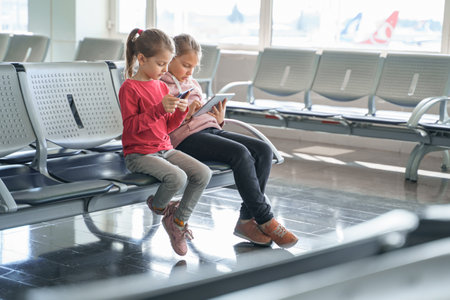 Children, kids, girls waiting for plane flight in departure hall. Sitting on chairs, playing computer games in gadgets, mobile phone, tablet. Chatting in social networks, what video. study online.の写真素材