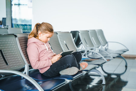 Kid, teen girl waiting for plane flight in departure hall. Sitting on chairs, playing computer games in gadgets, mobile phone, tablet. Chatting in social networks, what video. study online.の写真素材