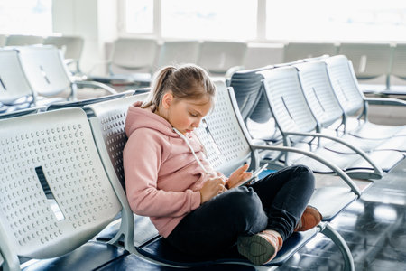 Kid, teen girl waiting for plane flight in departure hall. Sitting on chairs, playing computer games in gadgets, mobile phone, tablet. Chatting in social networks, what video. study online.の写真素材