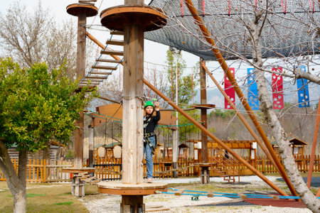 Teenage teen girl in climbing harness equipment, green sports safety helmet. Rope amusement park. Fastening attaching carabiner to safety rope. Hanging obstacle course. Kids children's attraction.の写真素材