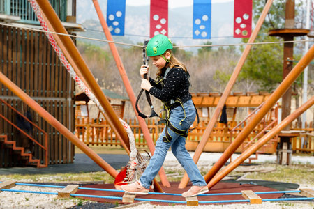 Teenage teen girl in climbing harness equipment, green sports safety helmet. Rope amusement park. Fastening attaching carabiner to safety rope. Hanging obstacle course. Kids children's attraction.の写真素材