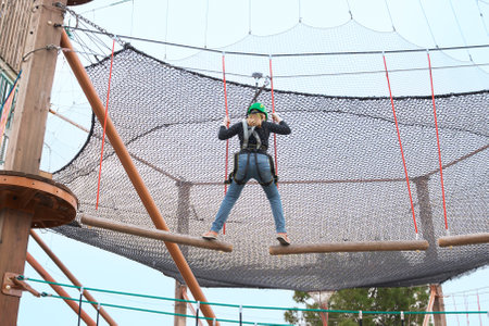 Teenage teen girl in climbing harness equipment, green sports safety helmet. Rope amusement park. Fastening attaching carabiner to safety rope. Hanging obstacle course. Kids children's attraction.の写真素材