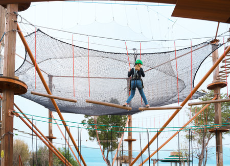 Teenage teen girl in climbing harness equipment, green sports safety helmet. Rope amusement park. Fastening attaching carabiner to safety rope. Hanging obstacle course. Kids children's attraction.の写真素材