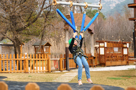 Teenage teen girl in climbing harness equipment, green sports safety helmet. Rope amusement park. Fastening attaching carabiner to safety rope. Hanging obstacle course. Kids children's attraction.の写真素材