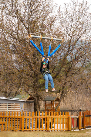 Teenage teen girl in climbing harness equipment, green sports safety helmet. Rope amusement park. Fastening attaching carabiner to safety rope. Hanging obstacle course. Kids children's attraction.の写真素材