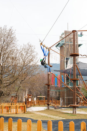 Teenage teen girl in climbing harness equipment, green sports safety helmet. Rope amusement park. Fastening attaching carabiner to safety rope. Hanging obstacle course. Kids children's attraction.の写真素材
