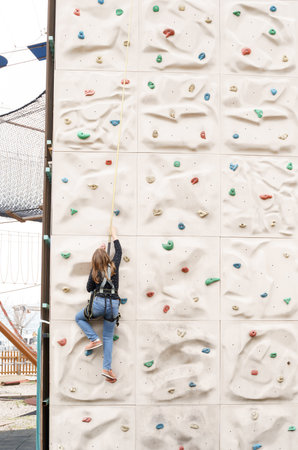 Teenage teen girl grabbing artificial high climbing wall with split ledges. Climbing harness safety sports equipment. Rope amusement city park, obstacle course. Kids children's attraction center.の写真素材