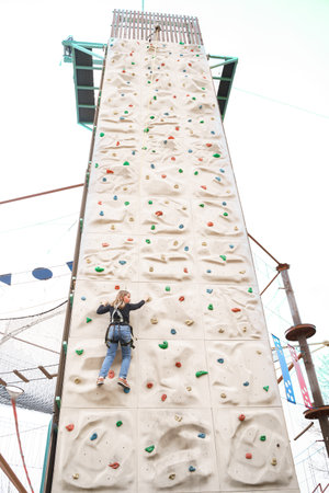 Teenage teen girl grabbing artificial high climbing wall with split ledges. Climbing harness safety sports equipment. Rope amusement city park, obstacle course. Kids children's attraction center.の写真素材