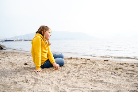 Lonely beautiful sad girl teenager sits thoughtfully on sand sea beach. Dreams, anxiety, worries about the future, school friends, parents. Teen bullying, psychological problems in adolescent puberty period.の写真素材