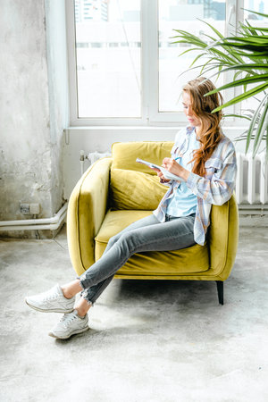 Young beautiful long haired college student girl in casual shirt with notebook,organizer pen sitting on yellow chair. Businesswoman making notes. Freelancer,remote job. Records in psychological diary.の写真素材