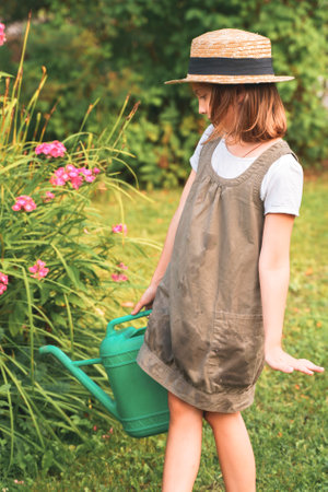 Farmer girl in summer straw hat. Little gardener farming, watering flowerbed with pink flowers, having fun in garden. Big green watering can water. Harvest help work. Cultivation plants, countryside.の写真素材