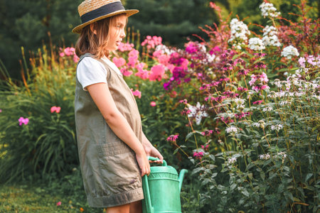 Farmer girl in summer straw hat. Little gardener farming, watering flowerbed with pink flowers, having fun in garden. Big green watering can water. Harvest help work. Cultivation plants, countryside.の写真素材