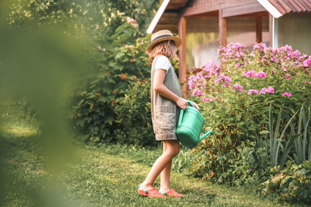 Farmer girl in summer straw hat. Little gardener farming, watering flowerbed with pink flowers, having fun in garden. Big green watering can water. Harvest help work. Cultivation plants, countryside.の写真素材
