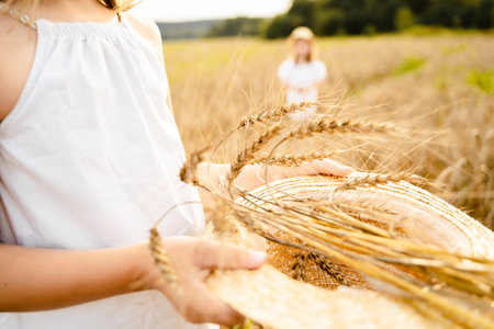 Happy girl in white dress with straw hat full of ears of wheat, rye, barley walking in yellow, orange field. Summer's yellow sun, nature freedom outdoors. Autumn harvest time rural scene. Own land.の写真素材