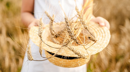 Happy girl in white dress with straw hat full of ears of wheat, rye, barley walking in yellow, orange field. Summer's yellow sun, nature freedom outdoors. Autumn harvest time rural scene. Own land.の写真素材