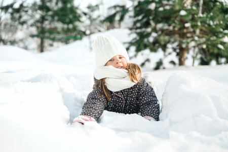 Little cute girl in white hat, scarf snood is lying, playing in snow. Kid walking in beautiful frozen forest, park among fir snowy trees. Winter vacation entertainment in village country house backyard.の写真素材