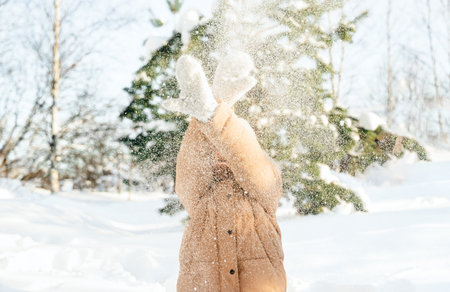 Little cute girl in white hat, scarf snood is lying, playing in snow. Kid walking in beautiful frozen forest, park among fir snowy trees. Winter vacation entertainment in village country house backyard.の写真素材