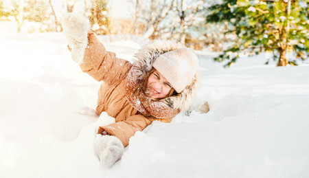 Little cute girl in beige hat,orange scarf snood is lying,playing in snow. Kid walking in beautiful frozen forest park among snowy trees. Winter vacation entertainment. Village country house backyard.の写真素材