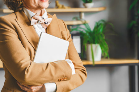 Confident stylish mature middle aged elderly woman in elegant suit, white shirt, bow tie with tablet...