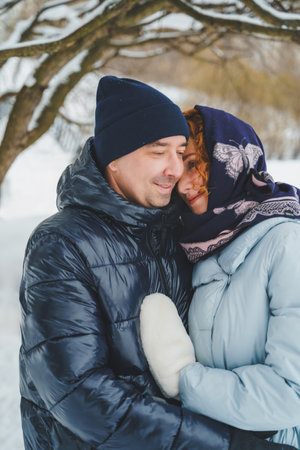 Love romantic love story. Happy couple of middle aged millennials, bright red-haired woman in winter snow park. Romantic date, kissing, hugging. Walking, having fun. Stylish blue clothes, jacket, hat.の写真素材