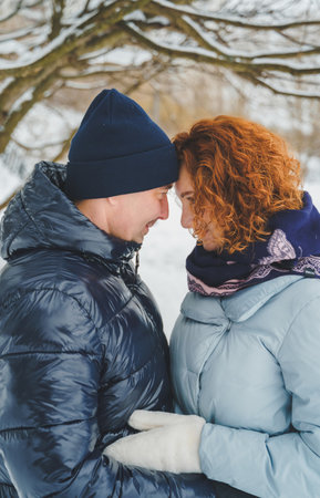 Love romantic love story. Happy couple of middle aged millennials, bright red-haired woman in winter snow park. Romantic date, kissing, hugging. Walking, having fun. Stylish blue clothes, jacket, hat.の写真素材