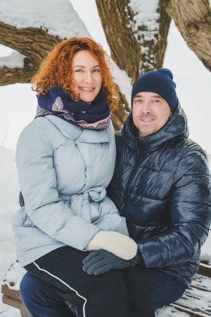 Love romantic love story. Happy couple of middle aged millennials, bright red-haired woman in winter snow park. Romantic date, kissing, hugging. Walking, having fun. Stylish blue clothes, jacket, hat.の写真素材