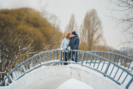 Love romantic love story. Happy couple of middle aged millennials, bright red-haired woman in winter snow park. Romantic date, kissing, hugging. Walking, having fun. Stylish blue clothes, jacket, hat.の写真素材