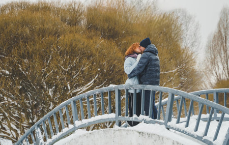 Love romantic love story. Happy couple of middle aged millennials, bright red-haired woman in winter snow park. Romantic date, kissing, hugging. Walking, having fun. Stylish blue clothes, jacket, hat.の写真素材