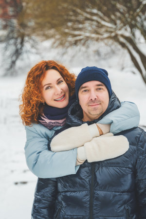 Love romantic love story. Happy couple of middle aged millennials, bright red-haired woman in winter snow park. Romantic date, kissing, hugging. Walking, having fun. Stylish blue clothes, jacket, hat.の写真素材