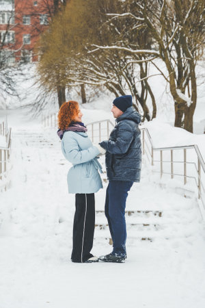 Love romantic love story. Happy couple of middle aged millennials, bright red-haired woman in winter snow park. Romantic date, kissing, hugging. Walking, having fun. Stylish blue clothes, jacket, hat.の写真素材