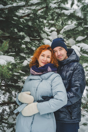 Love romantic love story. Happy couple of middle aged millennials, bright red-haired woman in winter snow park. Romantic date, kissing, hugging. Walking, having fun. Stylish blue clothes, jacket, hat.の写真素材