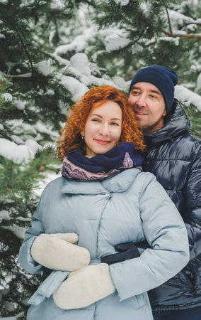 Love romantic love story. Happy couple of middle aged millennials, bright red-haired woman in winter snow park. Romantic date, kissing, hugging. Walking, having fun. Stylish blue clothes, jacket, hat.の写真素材