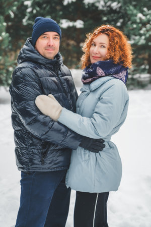 Love romantic love story. Happy couple of middle aged millennials, bright red-haired woman in winter snow park. Romantic date, kissing, hugging. Walking, having fun. Stylish blue clothes, jacket, hat.の写真素材