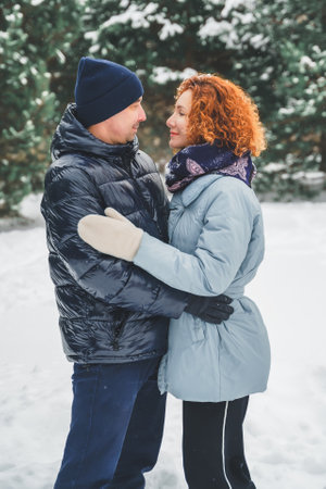 Love romantic love story. Happy couple of middle aged millennials, bright red-haired woman in winter snow park. Romantic date, kissing, hugging. Walking, having fun. Stylish blue clothes, jacket, hat.の写真素材