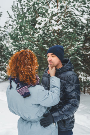 Love romantic love story. Happy couple of middle aged millennials, bright red-haired woman in winter snow park. Romantic date, kissing, hugging. Walking, having fun. Stylish blue clothes, jacket, hat.の写真素材