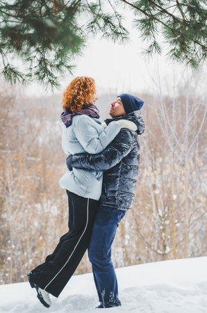 Love romantic love story. Happy couple of middle aged millennials, bright red-haired woman in winter snow park. Romantic date, kissing, hugging. Walking, having fun. Stylish blue clothes, jacket, hat.の写真素材