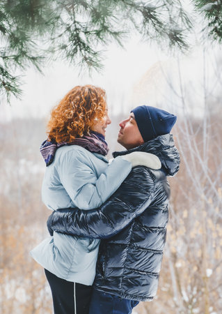 Love romantic love story. Happy couple of middle aged millennials, bright red-haired woman in winter snow park. Romantic date, kissing, hugging. Walking, having fun. Stylish blue clothes, jacket, hat.の写真素材