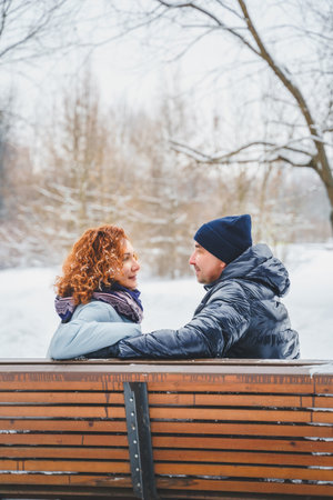 Love romantic love story. Happy couple of middle aged millennials, bright red-haired woman in winter snow park. Romantic date, kissing, hugging. Walking, having fun. Stylish blue clothes, jacket, hat.の写真素材